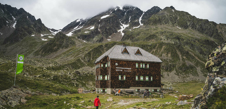 Ein Mann mit roter Jacke steht vor der Hochschober Huette an einem bewölkten Tag.