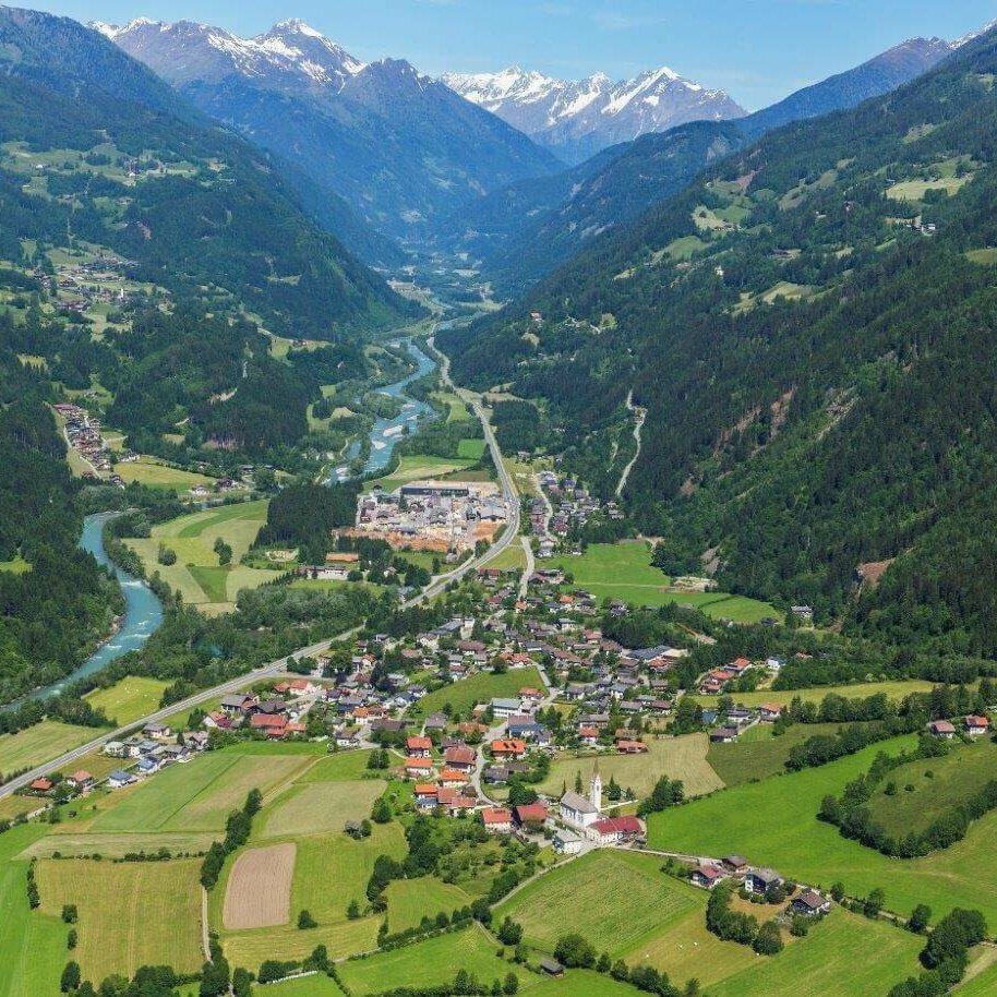 Luftaufnahme von Ainet im Sommer mit den grünen Wiesen und den Waldhängen. Im Hintergrund leuchten von den Bergen noch einige Schneefelder herunter.