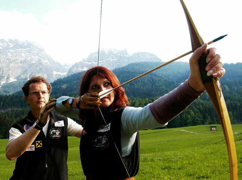 Ein Mann erklärt einer Frau die richtige Benützung eines Bogens. Die Frau hat den gespannten Bogen vor sich und zielt.