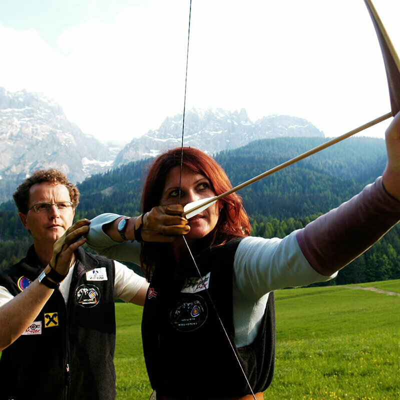 Ein Mann erklärt einer Frau die richtige Benützung eines Bogens. Die Frau hat den gespannten Bogen vor sich und zielt.