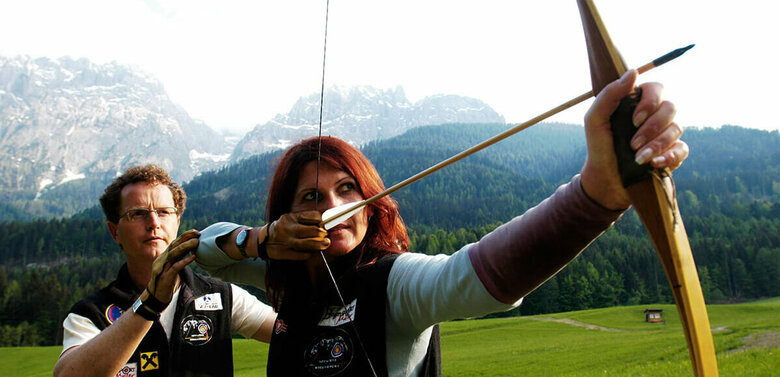 Ein Mann erklärt einer Frau die richtige Benützung eines Bogens. Die Frau hat den gespannten Bogen vor sich und zielt.