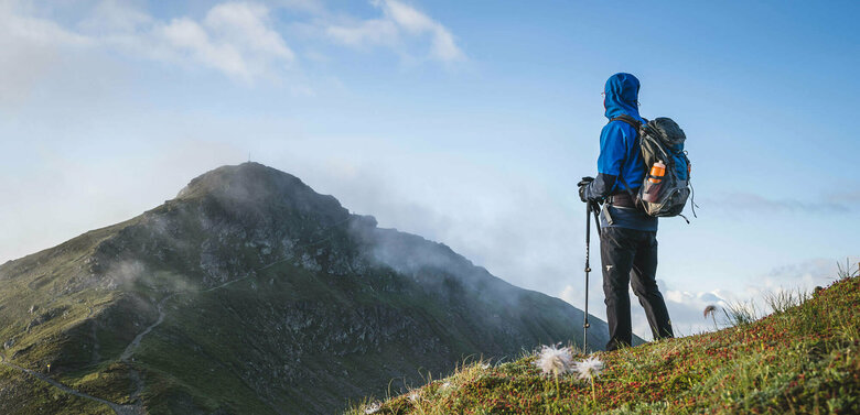 Schöne, frische Morgenstimmung - ein gut eingepackter Wanderer mit langer, dunkelgrauer Berghose, blauem Anorak mit übergezogener Kapuzze und blau-grauem Rucksack und angesteckter heller Trinkflasche blickt zu dem in einiger Entfernung liegenden Bergziel, das sich noch vom morgendlichen, leichten Nebel befreit.