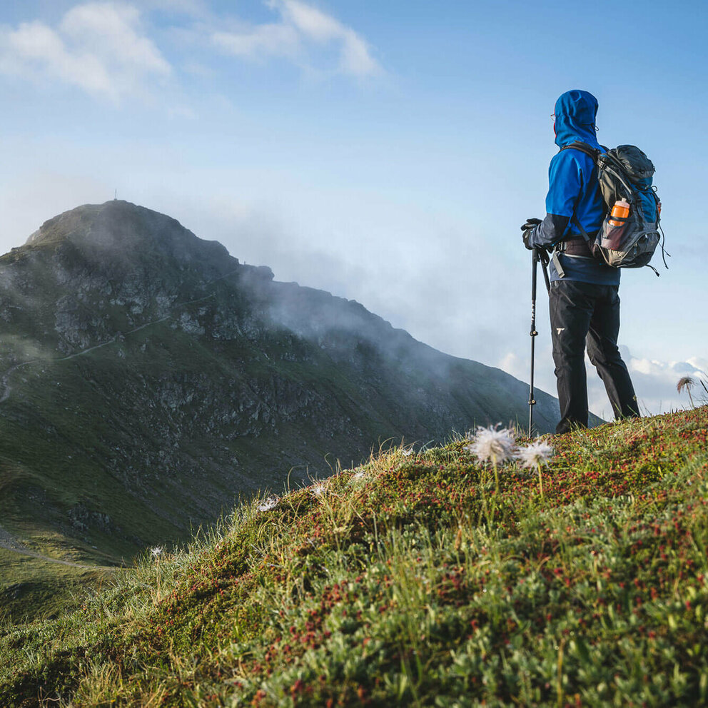 Schöne, frische Morgenstimmung - ein gut eingepackter Wanderer mit langer, dunkelgrauer Berghose, blauem Anorak mit übergezogener Kapuzze und blau-grauem Rucksack und angesteckter heller Trinkflasche blickt zu dem in einiger Entfernung liegenden Bergziel, das sich noch vom morgendlichen, leichten Nebel befreit.
