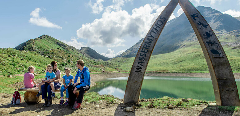 Wassermythos Ochsenlacke St. Jakob Brunnalm Eine Familie sitzt auf einer Bank am Ufer des Wassermythos Ochsenlacke im Skizentrum St. Jakob i. D.. Im Hintergrund sieht man die dazugehörigen Spielgeräte und das umliegende Bergpanorama, welches durch das schöne Wetter gut zur Geltung kommt.