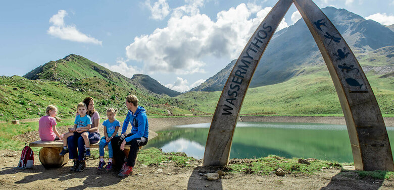 Eine Familie sitzt auf einer Bank am Ufer des Wassermythos Ochsenlacke im Skizentrum St. Jakob i. D..  Im Hintergrund sieht man die dazugehörigen Spielgeräte und das umliegende Bergpanorama, welches durch das schöne Wetter gut zur Geltung kommt.