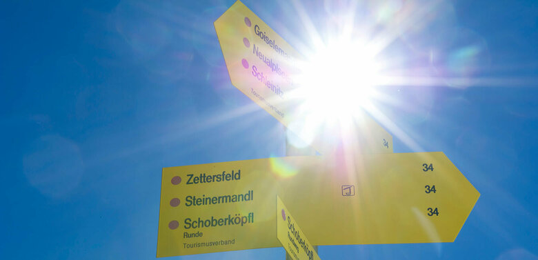 Ein gelber Wegweiser zur Orientierung am Zettersfeld steht von der Sonne geblendet im blauen Himmel.
