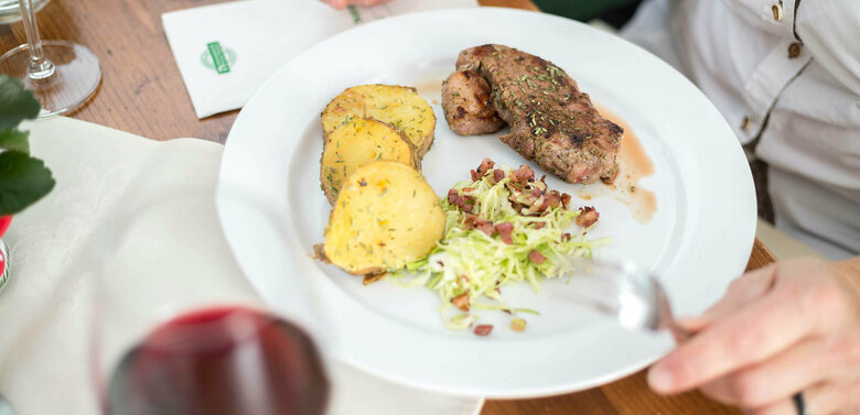 Gebratenes Fleischstück mit dicken Kartoffelscheiben und Krautsalat mit Speckwürfeln schön auf einem weißen Teller angerichtet mit einem Glas Rotwein.