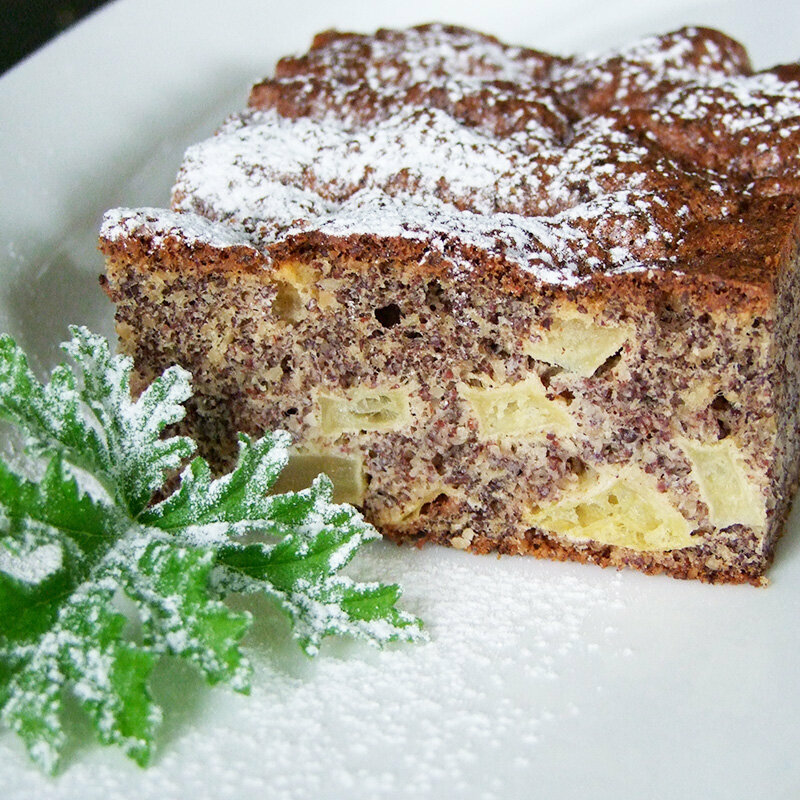 Ein Apfel-Mohnkuchen auf einem Teller, mit einem grünen Blatt und Puderzucker dekoriert.