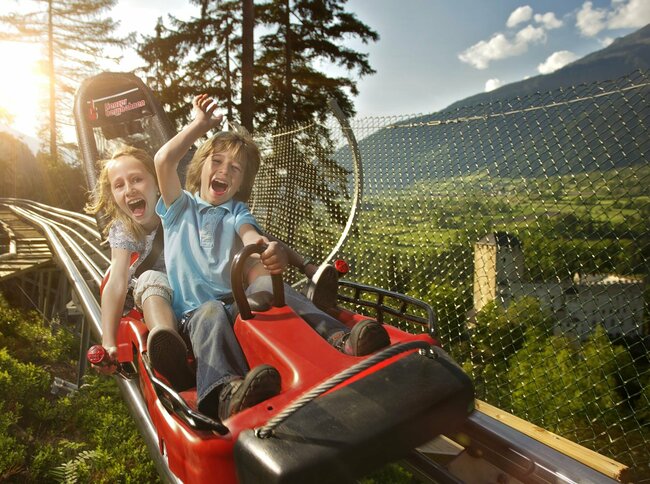 Ganzjahresrodelbahn Osttirodler am Hochstein Zwei Kinder fahren lachend mit der roten Sommerrodelbahn in Lienz in Osttirol. Im Hintergrund steht Schloss Bruck.