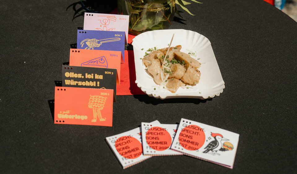 Auf einem Stehtisch mit schwarzer Tischdecke liegen die verschiedenen Gutscheine des Gluschtspechts. Daneben steht eine Portion geröstete Schlipfkrapfen und ein kleines Blumensträußchen.