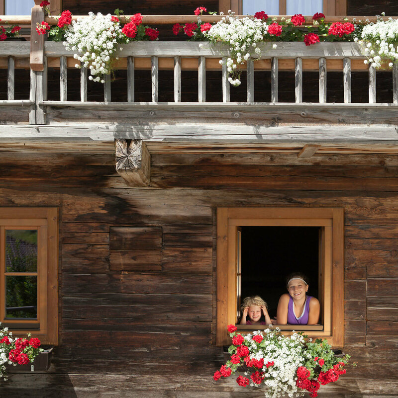 Zwei Kinder schauen aus dem Fenster eines Bauernhofs. An den Balkonen sind weiße und rote Blumen.