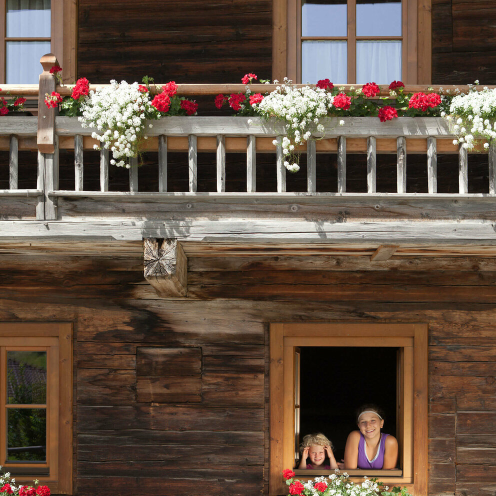 Zwei Kinder schauen aus dem Fenster eines Bauernhofs. An den Balkonen sind weiße und rote Blumen.