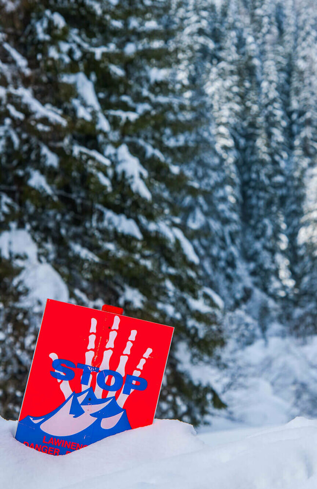 Eine rot, weiß, blaue Lawinengefahrtafel in einem frisch verschneitem Wald.