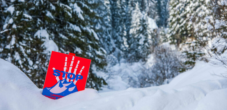 Eine rot, weiß, blaue Lawinengefahrtafel in einem frisch verschneitem Wald.