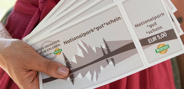 Nationalpark Gutscheine Frau im Dirndl zeigt mehrere Nationalpark Gutscheine im Wert von 5 Euro.