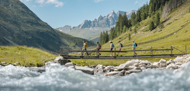 Vier Personen gehen hinter einem Nationalpark Ranger über eine kleine Holzbrücke, mit beeindruckender Bergkulisse im Hintergrund.