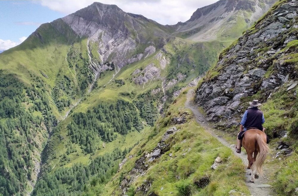 Ein Mann mit einem Hut reitet auf einem braunen Pferd auf einem schmalen Steig auf Muhs Panoramaweg.