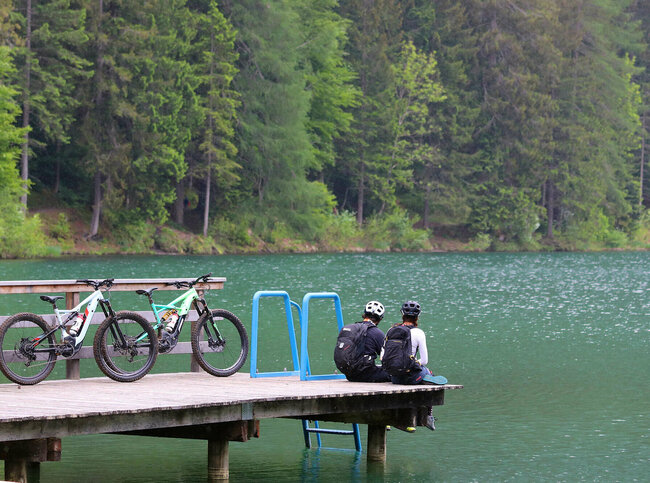 Ein Pärchen sitzt auf dem Holzsteg am Tristacher See und blickt aufs Wasser. Hinter ihenen stehen ihre E-Bikes.