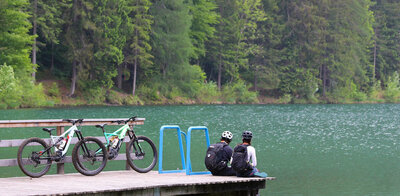 Ein Pärchen sitzt auf dem Holzsteg am Tristacher See und blickt aufs Wasser. Hinter ihenen stehen ihre E-Bikes.