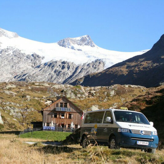 Ein blau-weißer Taxi-Bus vor einem Holzhaus mit schneebedeckten Bergen im Hintergrund.