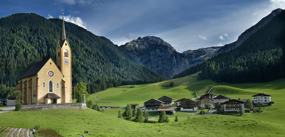Kartitsch Zwei Radfahrer auf der Straße durch Kartitsch mit der Pfarrkirche und den bewaldeten Hängen der Karnischen Alpen im Sommer im Hintergrund.