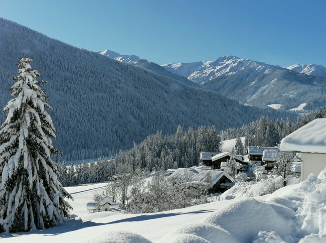 Kleine Häuseransammlung des Ortes Abfaltersbach umgeben von schneebedeckten Feldern und ein paar frisch verschneiten Bäume. Im Hintergrund tief winterliche, bewaldete Hänge und schneebedeckte Gipfel.