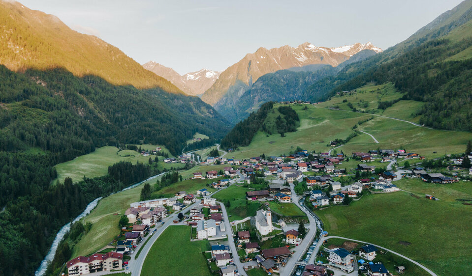 Blick von oben auf das Dorf Prägraten mit der Kirche im Zentrum im Frühling. Die Sonne scheint auf die Berge im Hintergrund. Links fließt ein kleiner Bach neben dem Dorf.
