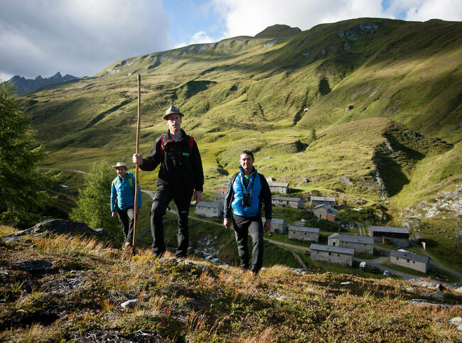 Drei Erwachsene wandern durch Umgebung der Jagdhausalmen. Im Hintergrund stehen mehrere alte Gebäude der Jagdhausalmen.