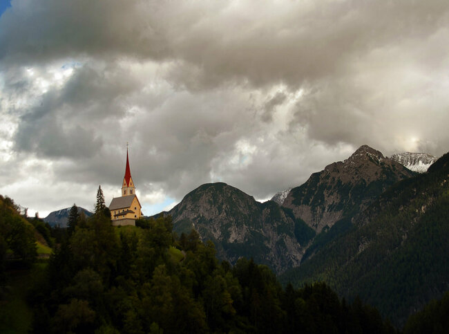 Kirche St. Justina inmitten eines Hanges umgeben von Bäumen und den Lienzer Dolomiten im Hintergrund.