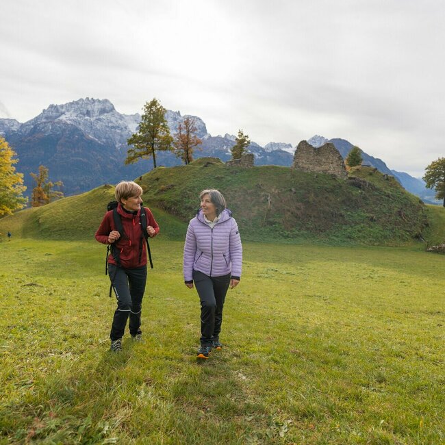 Zwei Damen wandern über eine grüne Wiese und lächeln sich zu. Im Hintergrund sieht man die Ruine Walchenstein.