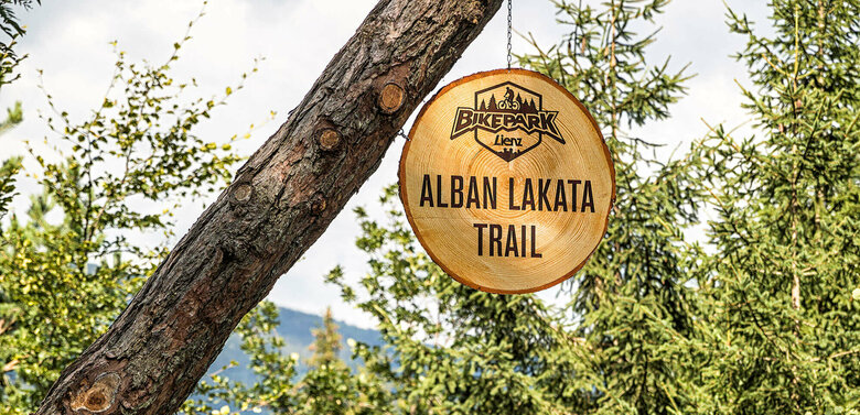 Zwei Radfahrer mit roter Bekleidung und gelben Helmen am Eingang des Alban Lakata Trails im Bikepark Lienz.