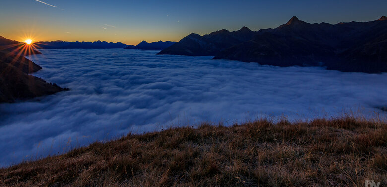 Sonnenaufgang über einem Wolkenmeer in einer weiten Berglandschaft. Einige Flugzeugen durchkreuzen den tiefblauen Himmel.