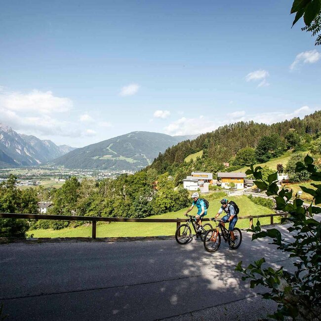 Zwei Radfahren fahren eine asphaltierte Straße bergauf, mit Blick auf Nussdorf Debant und den Lienzer Talboden.