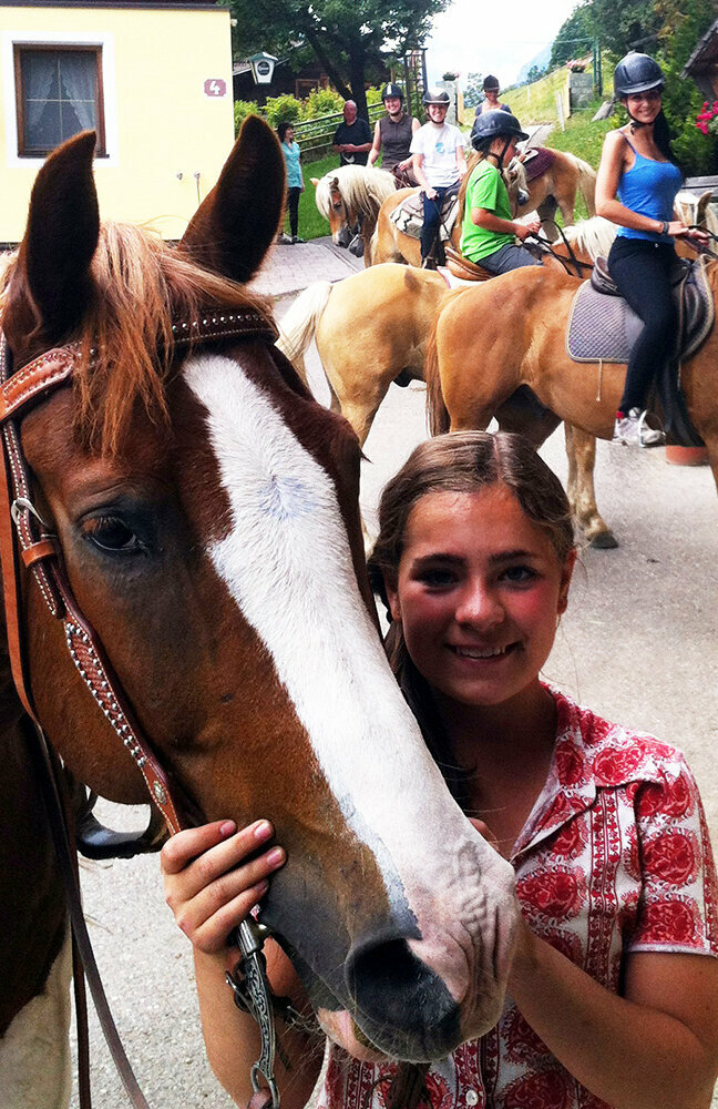 Reitbauernhof Ein Mädchen steht neben einem Pferd mit Trense auf einem Reitbauernhof. Im Hintergrund sind weitere Personen auf Pferden.