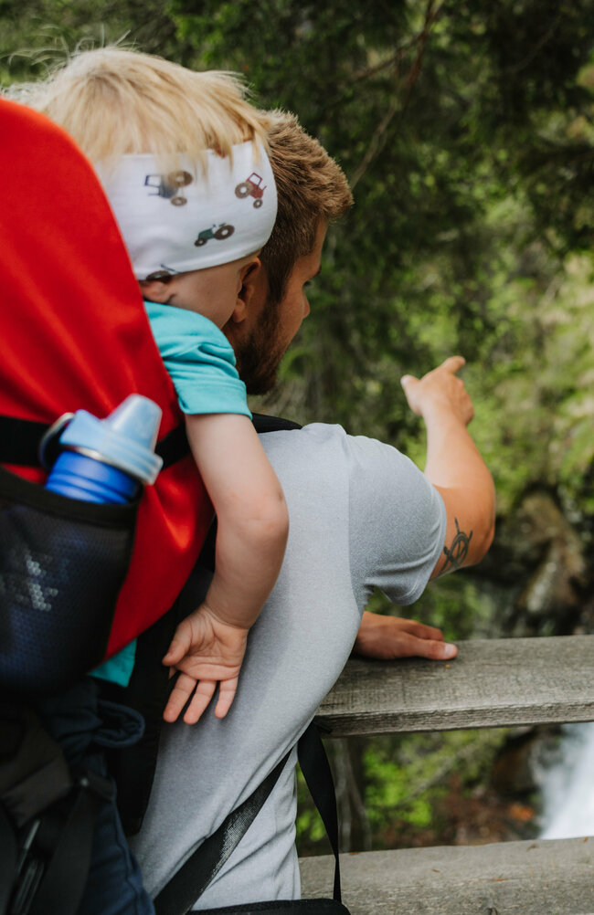 Ein Mann mit einem kleinen Kind auf dem Rücken zeigt einem Mädchen den Wasserfall im Kristeinertal.