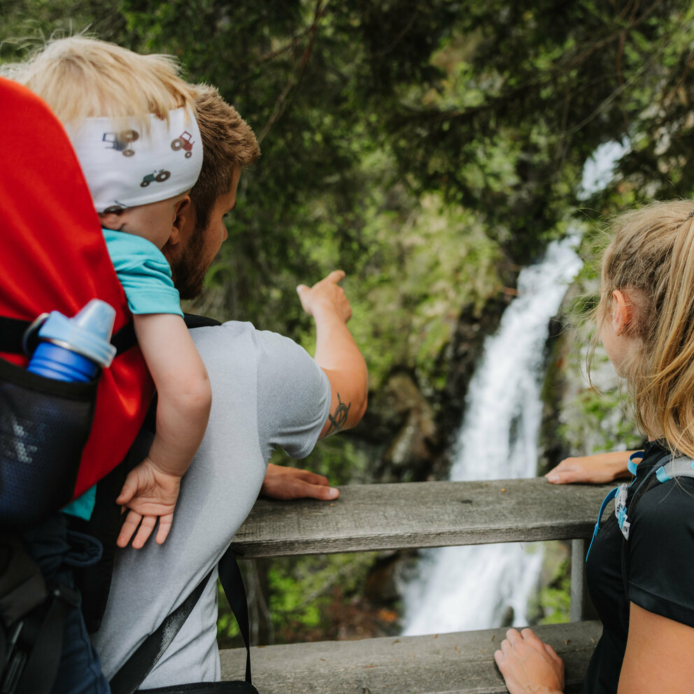 Ein Mann mit einem kleinen Kind auf dem Rücken zeigt einem Mädchen den Wasserfall im Kristeinertal.