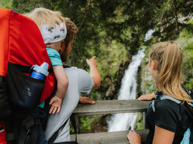 Ein Mann mit einem kleinen Kind auf dem Rücken zeigt einem Mädchen den Wasserfall im Kristeinertal.