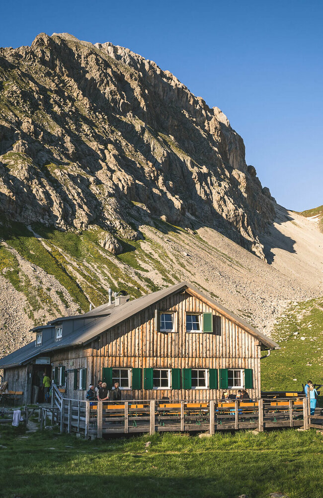 Obstanser Seehütte Ein rustikales Berghaus aus Holz mit grünen Fensterläden steht in einer sonnigen, alpinen Wiesenlandschaft. Im Hintergrund ragen steile, felsige Berggipfel auf, teils mit Schneefeldern bedeckt. Vor dem Haus sitzen und stehen einige Menschen, eine Holzterrasse mit Bänken lädt zum Verweilen ein