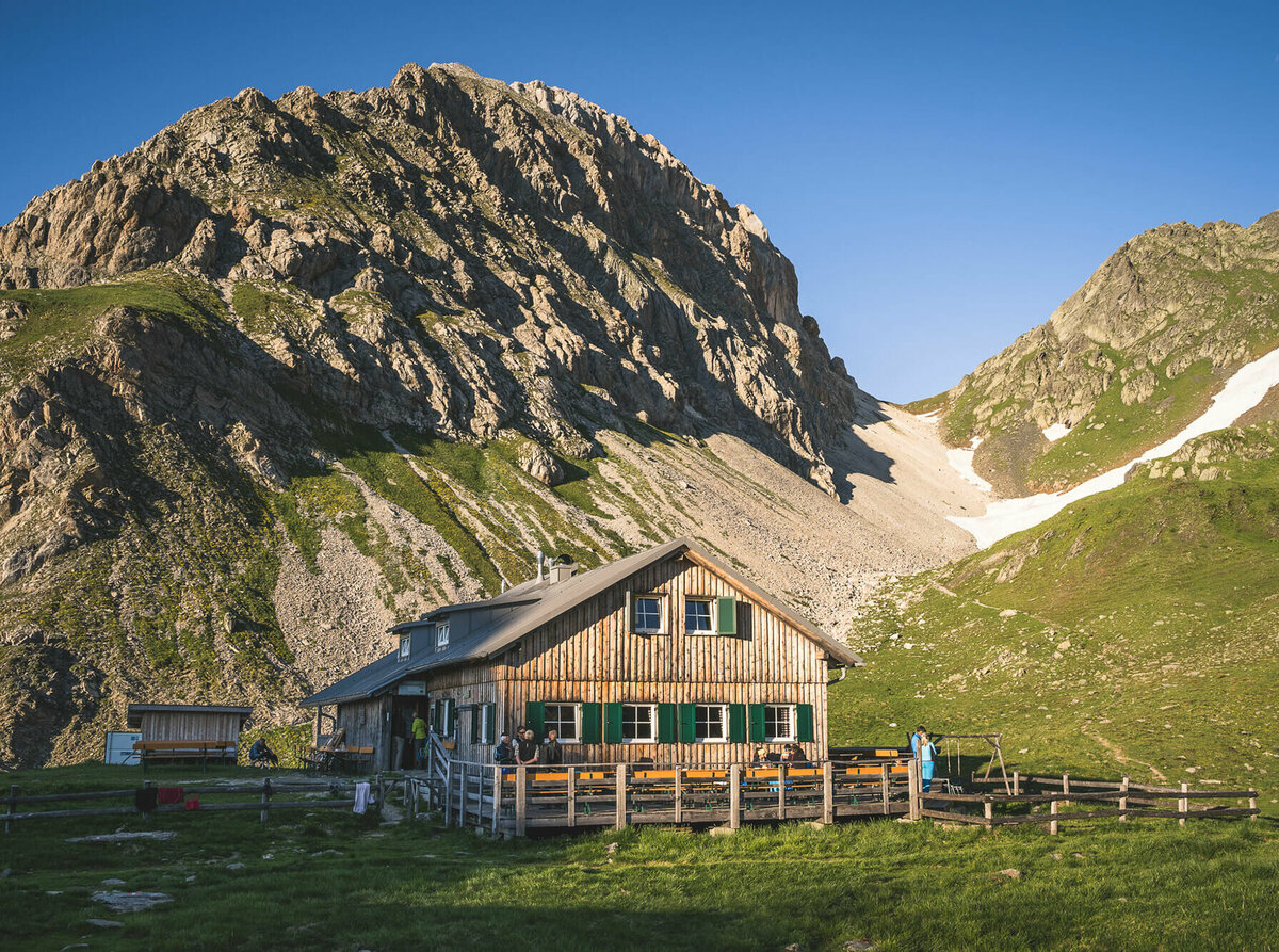 Ein rustikales Berghaus aus Holz mit grünen Fensterläden steht in einer sonnigen, alpinen Wiesenlandschaft. Im Hintergrund ragen steile, felsige Berggipfel auf, teils mit Schneefeldern bedeckt. Vor dem Haus sitzen und stehen einige Menschen, eine Holzterrasse mit Bänken lädt zum Verweilen ein