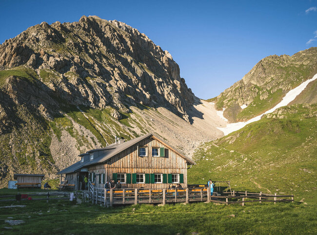 Obstanser Seehütte Ein rustikales Berghaus aus Holz mit grünen Fensterläden steht in einer sonnigen, alpinen Wiesenlandschaft. Im Hintergrund ragen steile, felsige Berggipfel auf, teils mit Schneefeldern bedeckt. Vor dem Haus sitzen und stehen einige Menschen, eine Holzterrasse mit Bänken lädt zum Verweilen ein
