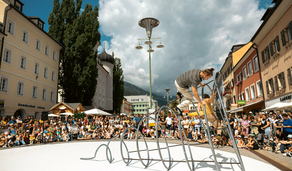 Zwei Akrobaten von The Tea Time Company turnen im Rahmen von Olala auf einer großen Spirale aus Eisen im Kreis am Lienzer Hauptplatz. Viele Schaulustige sind hier am Lienzer Hauptplatz, um sich das Spektakel anzusehen.