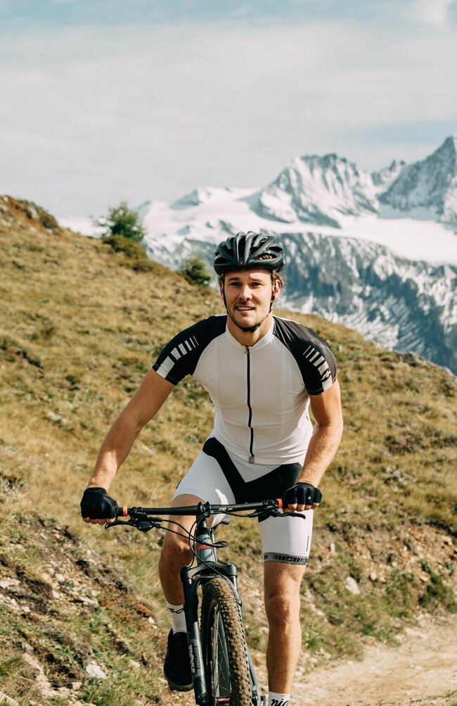 Ein Radfahrer von vorne, mit schwarz-weißer Radbekleidung und schwarzem Helm. Er fährt auf einem schmalen Trail im Bikepark Großglockner mit dem besagten Berg im Hintergrund.