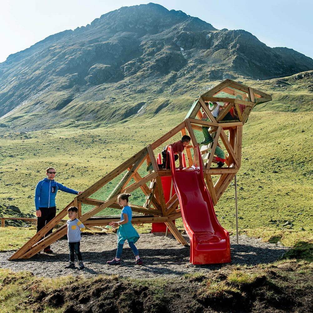 Zu sehen sind glückliche Kinder auf einem Spielgerät, welches einem überdimensional-großen Vogel ähnlich sieht, des Wassermythos Ochsenlacke im Skizentrum St. Jakob i. D.. Das sonnige Wetter lässt die umliegende Umgebung und die Berge im Hintergrund in einem warmen Licht erstrahlen.