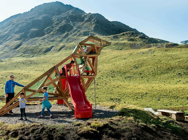 Zu sehen sind glückliche Kinder auf einem Spielgerät, welches einem überdimensional-großen Vogel ähnlich sieht, des Wassermythos Ochsenlacke im Skizentrum St. Jakob i. D.. Das sonnige Wetter lässt die umliegende Umgebung und die Berge im Hintergrund in einem warmen Licht erstrahlen.
