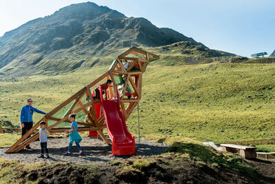 Wassermythos Ochsenlacke St. Jakob Brunnalm Zu sehen sind glückliche Kinder auf einem Spielgerät, welches einem überdimensional-großen Vogel ähnlich sieht, des Wassermythos Ochsenlacke im Skizentrum St. Jakob i. D.. Das sonnige Wetter lässt die umliegende Umgebung und die Berge im Hintergrund in einem warmen Licht erstrahlen.