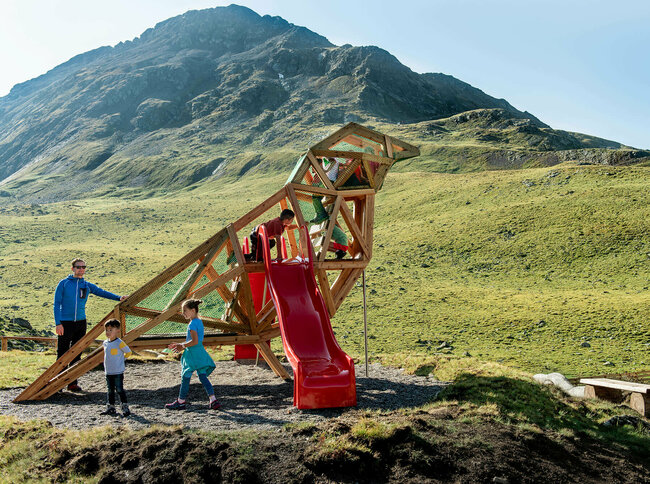 Wassermythos Ochsenlacke St. Jakob Brunnalm Zu sehen sind glückliche Kinder auf einem Spielgerät, welches einem überdimensional-großen Vogel ähnlich sieht, des Wassermythos Ochsenlacke im Skizentrum St. Jakob i. D.. Das sonnige Wetter lässt die umliegende Umgebung und die Berge im Hintergrund in einem warmen Licht erstrahlen.