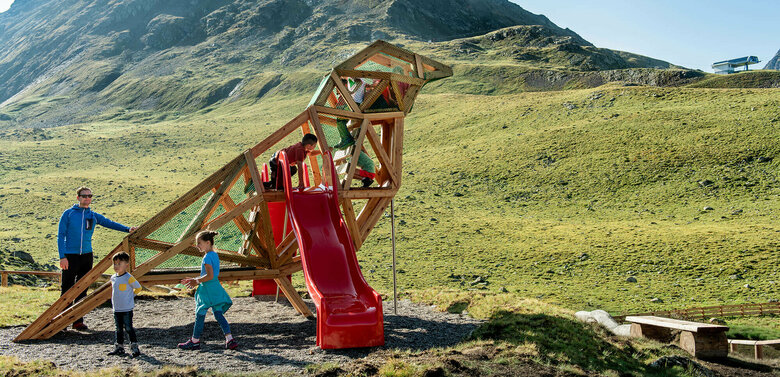 Zu sehen sind glückliche Kinder auf einem Spielgerät, welches einem überdimensional-großen Vogel ähnlich sieht, des Wassermythos Ochsenlacke im Skizentrum St. Jakob i. D.. Das sonnige Wetter lässt die umliegende Umgebung und die Berge im Hintergrund in einem warmen Licht erstrahlen.