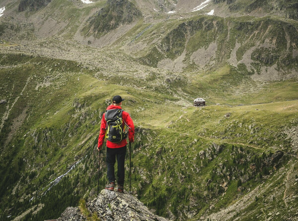Ein Wanderer mit roter Jacke und Rucksack steht auf einem Felsen mit Blick auf die Hochschoberhütte in Osttirol.