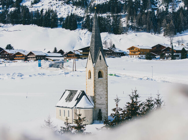 Die Kals Kirche mit dunklem Kirchturm liegt von Schnee bedeckt vor dem Dorf.