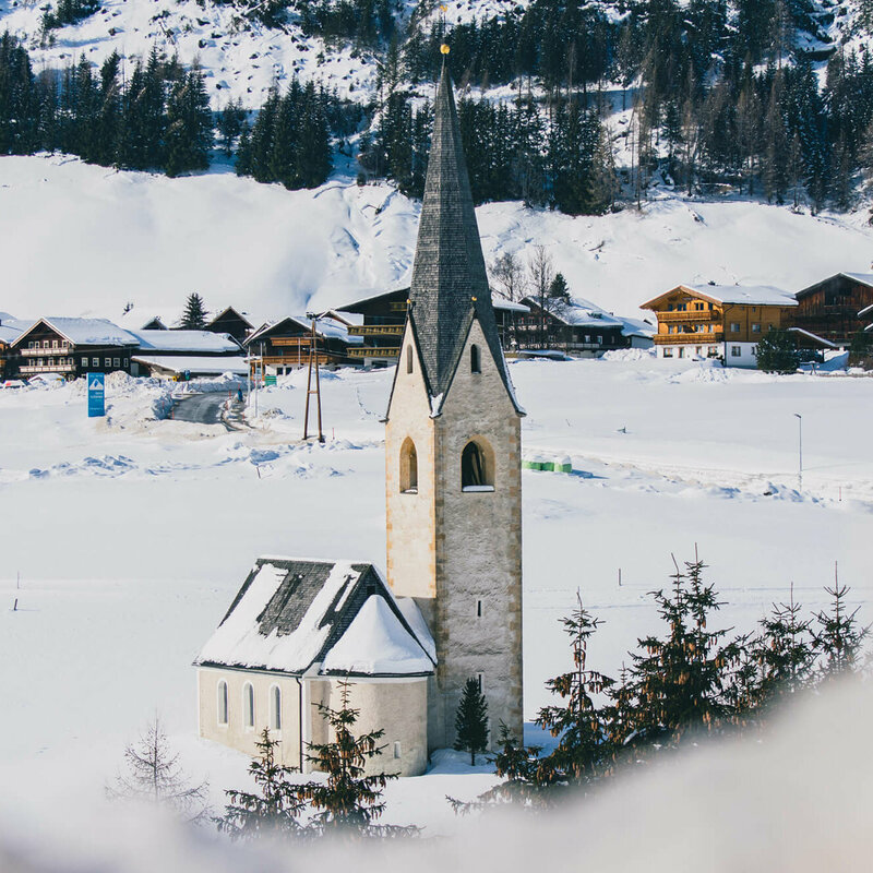 Die Kals Kirche mit dunklem Kirchturm liegt von Schnee bedeckt vor dem Dorf.